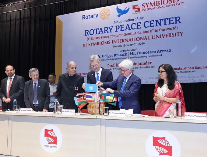 The image shows a group of individuals standing behind a table at an event for the inauguration of the Rotary Peace Center at Symbiosis International University. Various flags are displayed on the table, and banners with the Rotary and Symbiosis International University logos are visible in the background. One person is holding a globe symbolizing peace. The atmosphere is formal, with participants dressed in business attire.