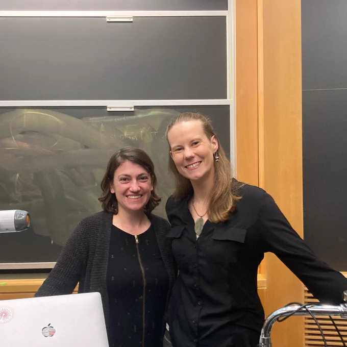Nicole Creanza and Kate Snyder stand in front of a chalkboard smiling while wearing black sweaters