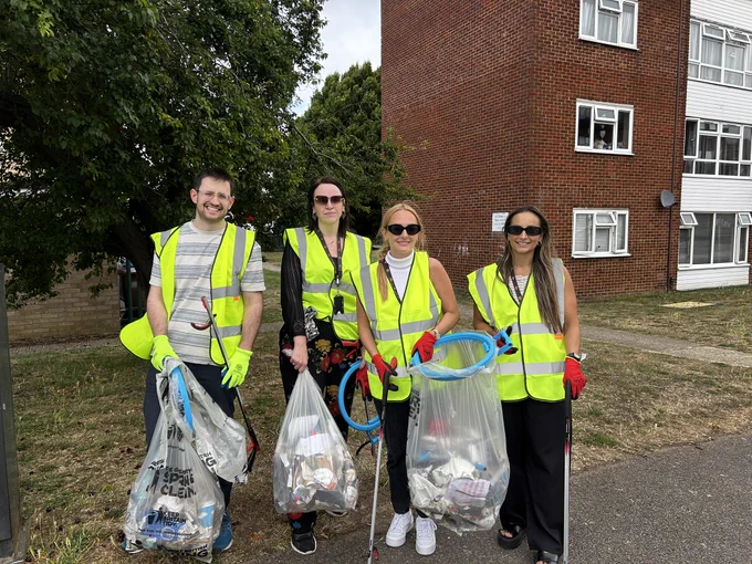 A group of four Settle colleagues wearing high‑visibility vests and gloves standing outdoors in a residential area. They are holding litter‑picking tools and large bags filled with collected rubbish. A flat block and a tree are visible in the background.