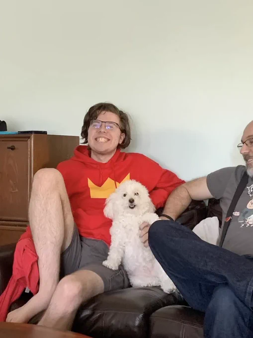 A picture of Technoblade sitting with his little white dog Floof. Floof is sitting up against Techno, with one paw on his leg. Techno is wearing a red hoodie and grey shorts. He's got a big smile! Technodad is mostly cropped out of the picture but he is also smiling and petting Floof. The picture is from "Floof Retrospective with Technoblade" from the Mister Technodad YouTube channel. 