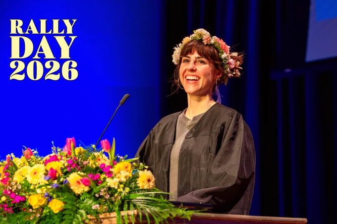 A person wearing a flower crown and graduation gown stands smiling behind a podium adorned with colorful flowers. The backdrop displays "Rally Day 2026" in bold yellow letters.