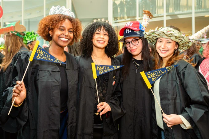 A group of four graduates in black gowns, each holding a small flag that says "Smith Seniors 2026." They are standing indoors, smiling, and wearing creative hats.