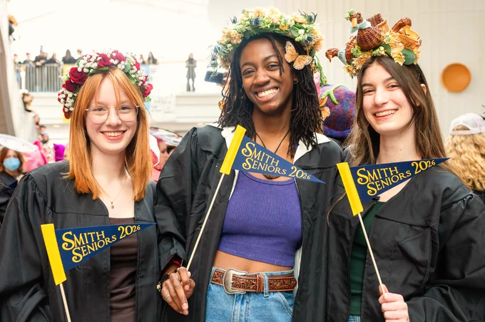 A group of three graduates in black gowns, each holding a small flag that says "Smith Seniors 2026." They are standing indoors, smiling, and wearing creative hats.