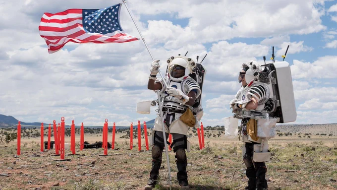 NASA astronaut Andre Douglas (left) raises an American flag next to NASA astronaut Kate Rubins (right) wearing partial spacesuits in a desert-like environment. 