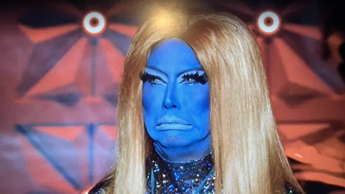 Serena Morena in full blueface with a long, straight orange wig, making a grumpy face in what is apparently the style of Crazy Frog.