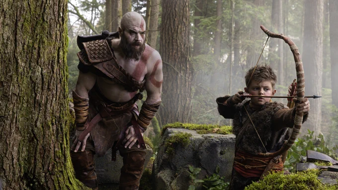 Large intimidating father with red tattoos watches his son dressed in furs prepare to shoot a bow and arrow. They are in a lush green forest. 