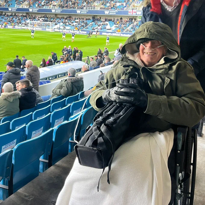 Eddie, 83, smiling at the camera at a football pitch, wrapped in a coat and blanket.
