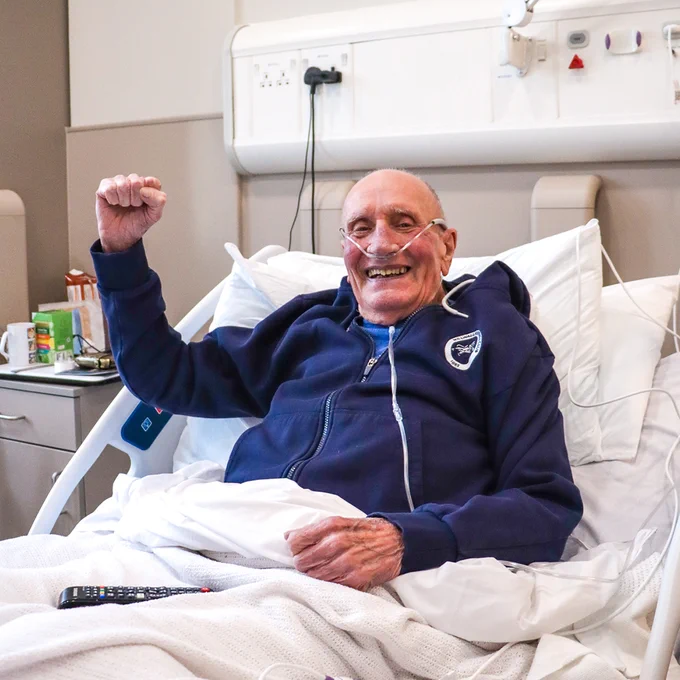 Eddie, 83, smiling in a hospice bed while wearing a Millwall jacket.