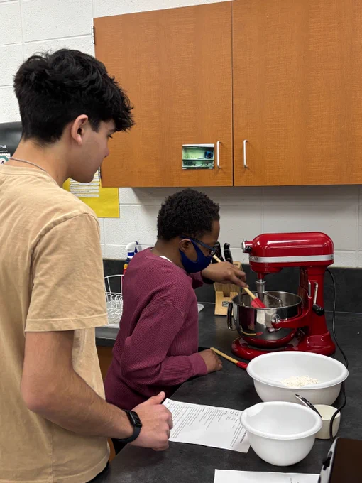 Two students working together in a kitchen, one stirring ingredients in a red stand mixer while the other reads a recipe sheet.