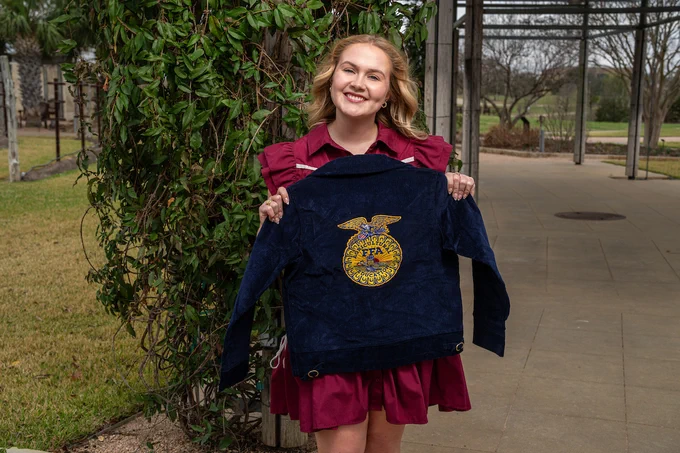 A person is outside holding a blue jacket with the FFA emblem. They are standing on a paved path surrounded by greenery.