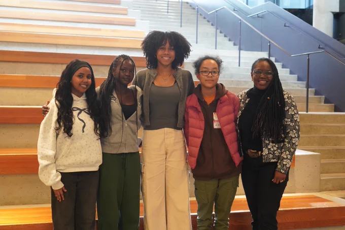 Group of five young adults standing together on wooden steps inside the UBC Education building with a staircase in the background.