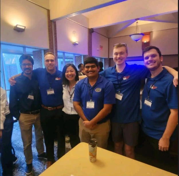 Kyle Groome, Michael Panarisi, and Hugo Resendez stands together with their colleagues indoors, some wearing blue polo shirts and badges. They are smiling and appear to be at an event or gathering.