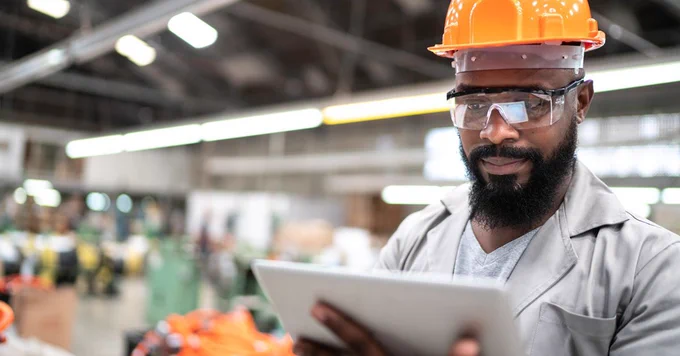 A person wearing a hard hat and safety glasses uses a tablet in an industrial setting.
