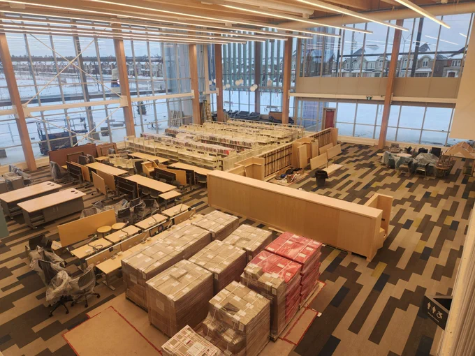 Large modern library interior under construction with stacks of boxed books and empty shelves near expansive windows overlooking a waterfront.