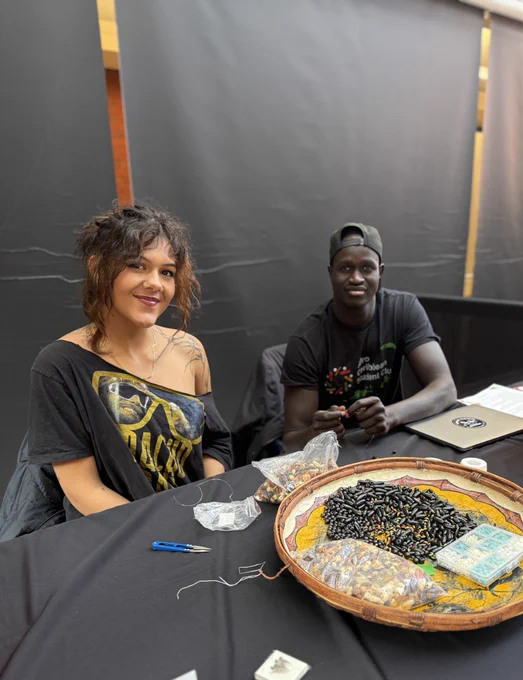 Two people are smiling and sitting at a table with supplies. The tabletop features a large, round tray filled with beads and necklaces. 
