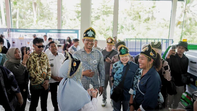 Suasana akrab dalam sebuah acara lingkungan di Kepulauan Derawan, Indonesia. Tampak beberapa pria dan wanita mengenakan penutup kepala tradisional berwarna hitam-emas dengan motif penyu. Seorang pria berbaju batik biru dan seorang wanita berhijab biru muda sedang tersenyum dan berinteraksi dengan warga serta pejabat setempat. Di latar belakang terlihat truk pengangkut sampah hijau dan banner program kebersihan.