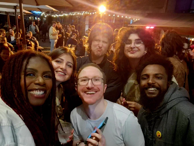6 people of various gender and race posing for a selfie in the outside seating area of a busy city bar at night. 