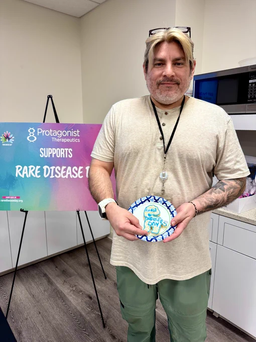 Protagonist holding their decorated zebra cookie in front of a colorful sign that says, "Protagonist Therapeutics supports Rare Disease Day"