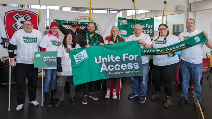 Rotherham United Disabled Supporters' Association members are joined by Level Playing Field's Owain Davies. Everyone is smiling towards camera, with 'Unite For Access' banners, boards, scarves and t-shirts.