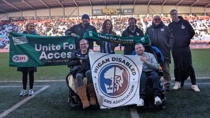 Members of the newly formed Wigan Disabled Supporters' Association are joined pitch side by Level Playing Field's Liam Bird. Everyone is smiling towards camera, with a 'Unite For Access' banner and scarf, while the two people at the front are holding a banner for the disabled supporters' association.