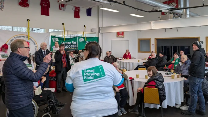 A room full of Rotherham United Disabled Supporters' Association members in discussion with one another. Some 'Unite For Access' banners are in the background, while some people are wearing t-shirts and scarves.