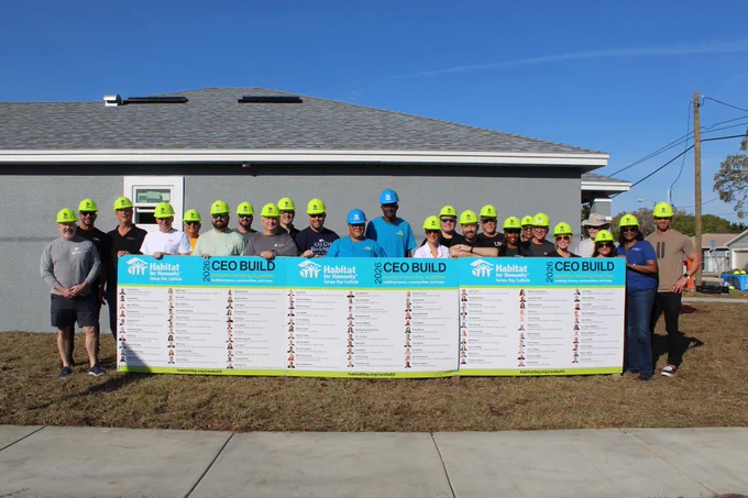 A group of people wearing yellow hard hats stand in front of a grey house. They are holding a large sign displaying the "Habitat for Humanity" logo and the words "CEO Build."