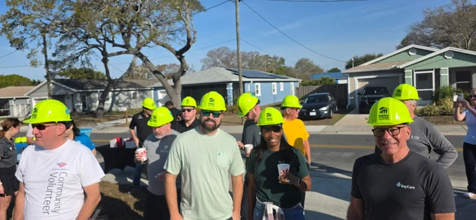 A group of people wearing bright green hard hats stand outdoors, participating in a community build event. Some of them are wearing shirts with logos, including Habitat for Humanity and BayCare. The scene includes residential houses and trees in the background.