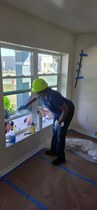 Dr. Tonjua Williams wearing a hard hat and working on a window from the inside. Two other individuals in hard hats are working outside the window. The room has preparations for painting, including plastic coverings on the floor.