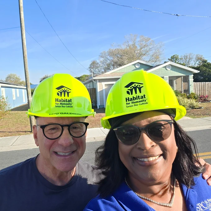 Two people, including Dr. Tonjua Williams, wearing bright yellow hard hats with "Habitat for Humanity" logos are smiling outdoors. They are positioned in front of a house on a sunny day.