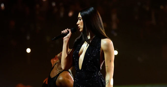 Dua Lipa is captured in profile while performing, holding a black microphone to her lips. She is wearing a plunging, sequined black bodysuit that sparkles under the stage lights, paired with fishnet stockings. Her long, dark hair is straight and a dancer is partially visible in the background against a dark arena.