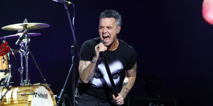 Robbie Williams performs live, captured with his mouth wide open in a powerful vocal moment. He is wearing a black t-shirt featuring a stylized graphic of eyes, dark trousers, and several silver rings and bracelets. His arms are covered in tattoos and his grey-toned hair is styled upwards.