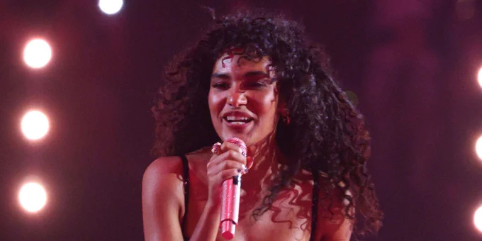 Olivia Dean captured mid-performance, singing into a glittery pink microphone. She has long, voluminous curly hair and is wearing a dark, sequinned dress that catches the purple and pink stage lights. 