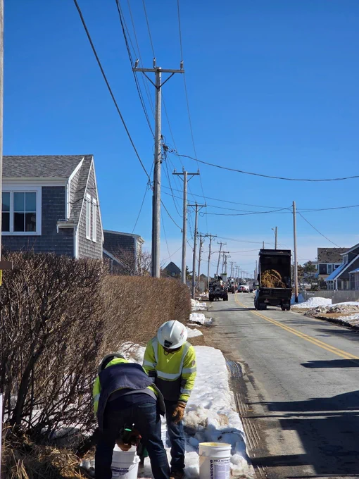 Two utility workers in safety gear kneel beside buckets on a snowy roadside, preparing materials as additional bucket trucks and crews work farther down the street. Homes and utility poles line the road beneath a bright blue sky.