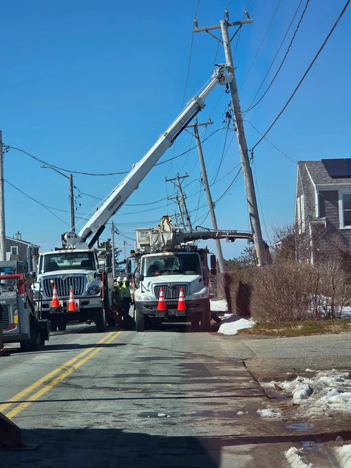 Bucket trucks and utility crews work on a leaning utility pole along a narrow residential road. Traffic cones line the front of the trucks as workers repair overhead wires under a clear blue sky.