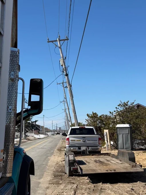 A utility pole leans sharply to one side while a pickup truck with a flatbed trailer is parked nearby. Snow patches, roadside brush, and overhead wires frame the scene on a sunny day.