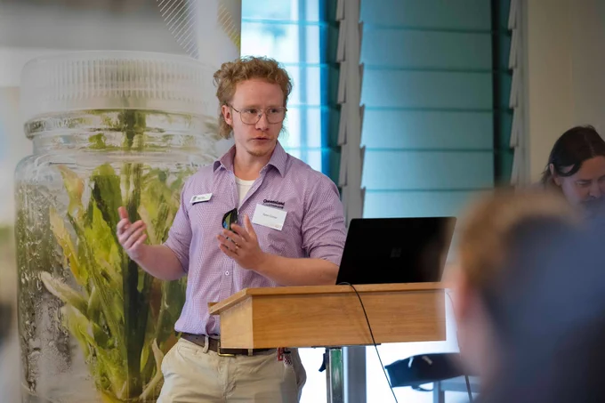 Man in a light purple shirt giving a presentation next to a wooden podium with a laptop in a room with blue window blinds at the AgriFutures Ginger research event and farm tour May 2025.