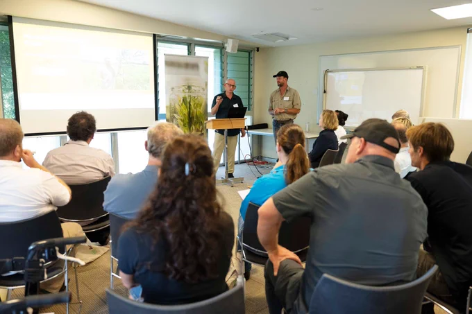 Two men presenting information to a seated audience in a bright conference room with a projector screen and whiteboard at the AgriFutures Ginger research event and farm tour May 2025.