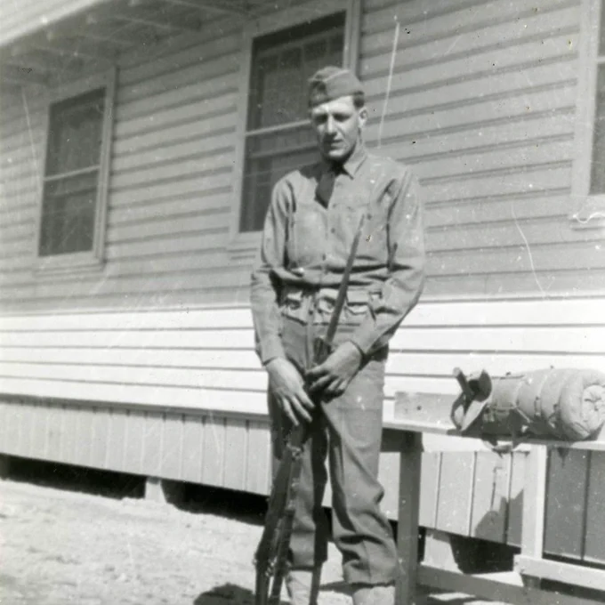 Historic black and white photo of a man in military uniform standing outside a paneled building on blocks holding a rifle vertically.