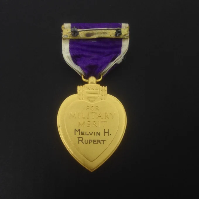 Gold heart-shaped medal with purple and white ribbon inscribed 'For Military Merit Melvin H. Rupert. Medal displayed against a black background.