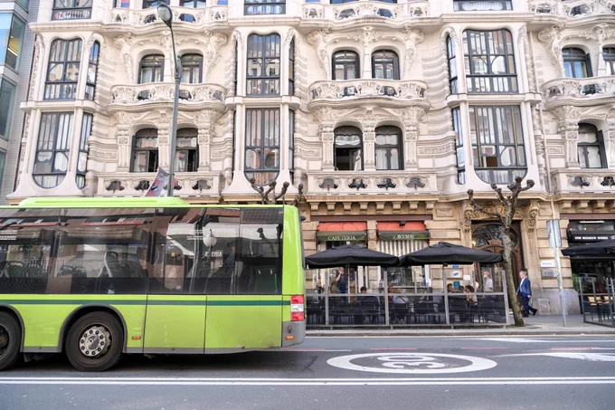 People of all ages and abilities walk, cycle, drive, and ride public transportation along the low-traffic streets of Bilbao, which are lined with buildings, trees, bike infrastructure, and prominent 30 km/h speed limit signage and road markings.