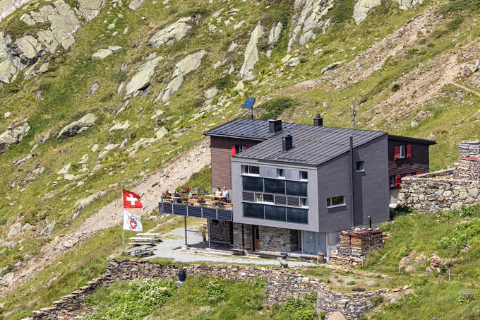 Die Silvrettahütte steht auf einem steilen, grünen Berghang. Das moderne Gebäude mit grauer Fassade und dunklem Dach hat eine Terrasse mit Tischen und Bänken. Daneben wehen eine Schweizer Fahne und eine SAC-Flagge. Im Hintergrund sind Felsen und alpine Wiesen zu sehen.