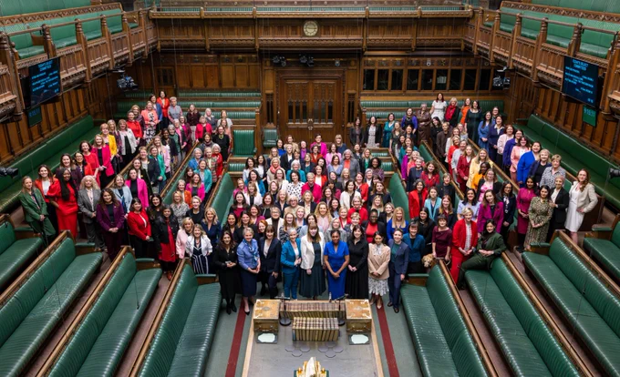 Women MPs standing together in the House of Commons Chamber, with green benches and wood-panelled walls.