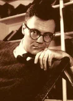 Lowell, with glasses and a sweater is sitting on a chair, exuding a relaxed and contemplative demeanor.