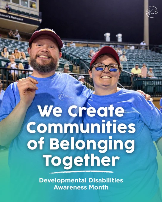 Two adults wearing blue SJCS T-shirts stand arm in arm at a baseball stadium, smiling widely. Stadium seats and spectators are visible behind them. Overlaid text reads: “We Create Communities of Belonging Together – Developmental Disabilities Awareness Month.”