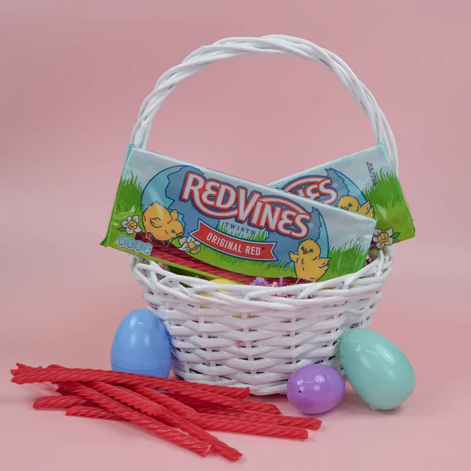 A white wicker basket filled with packages of Red Vines Original Red candy is set against a pink background. The basket also contains colorful plastic eggs. Five pieces of red licorice are arranged in front of the basket.