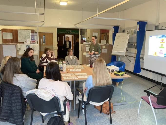 A group of people are seated around a table in a hospital setting, engaged in a discussion. One person is standing and speaking, while a presentation is displayed on a screen. The area is labeled "Emergency Department" at the back. The environment is informal and collaborative.