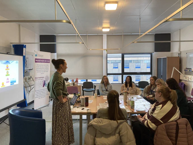A group of people is gathered in a meeting room. A person stands at the front presenting to others seated around a table. There's a projection screen displaying colorful graphics and a banner labeled "Mental Health Services." The room is well-lit with several windows.