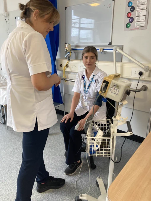 Two medical professionals in a hospital setting, one sitting and one standing, near medical equipment. Both are wearing uniforms, and the environment includes a whiteboard and medical devices.
