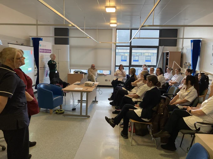 A group of people in a hospital setting are sitting and standing around a room, focused on a presentation. The attendees are dressed in medical uniforms, and banners with NHS and healthcare information are visible in the background.