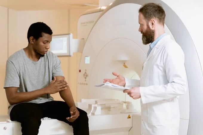 Foto van een denkbeeldig onderzoek. Man zit op een bank van een CT scan. Een man in witte jas met papieren in zijn hand spreekt hem toe. 